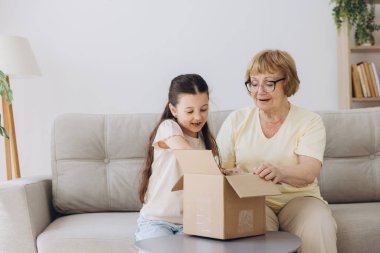 Karton kutuyu açan mutlu büyükanne ve torun kızı evdeki kanepede oturuyor. Şok olmuş aile tüketicileri iyi paketleri açıyor ve büyük alışverişin içine bakıyor..