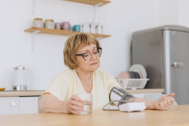 Yüksek tansiyonu olan yaşlı bir kadın evinde tansiyonunu ölçüyor. Üzgün yaşlı kadın kan basıncını tonometreyle ölçüyor. Günlük sağlık kontrolleri