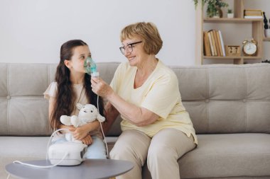 Küçük kız büyükannesiyle nebulizör soluyor. Kadın, hasta bir çocuğu kucaklarken soluk alıp veriyor. Büyükannem torununa soğuk algınlığı ve nezle konusunda yardım ediyor..