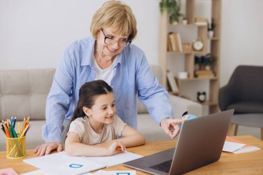 Mutlu büyükanne, kreş çocuğuna ev ödevi için yardım ediyor. Pozitif öğrenci kız ve büyükanne laptopta öğrenme kitabı okuyorlar, gülümsüyorlar, evde eğitimin tadını çıkarıyorlar.