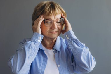 Evde baş ağrısı olan, acı çeken ve gri arka planda kendini iyi hissetmeyen yaşlı bir kadının portresi.