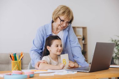 Mutlu büyükanne, kreş çocuğuna ev ödevi için yardım ediyor. Pozitif öğrenci kız ve büyükanne laptopta öğrenme kitabı okuyorlar, gülümsüyorlar, evde eğitimin tadını çıkarıyorlar.