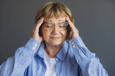 Evde baş ağrısı olan, acı çeken ve gri arka planda kendini iyi hissetmeyen yaşlı bir kadının portresi.