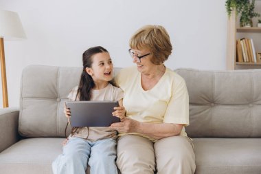 Eğleniyorum. Gülümseyen emekli kadın dadı rahat kanepeye oturup küçük kızı kucaklıyor internette tablet kullanarak komik çizgi filmler seyrediyor. Büyük anne ve küçük torun arasındaki bağ dijital bloknotta video klibinin keyfini çıkarın