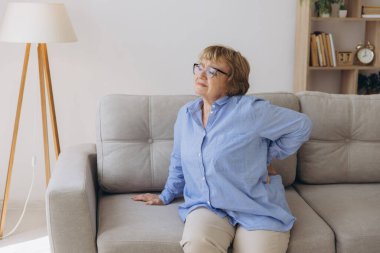 Mutsuz yaşlı bir kadın sırtına dokunuyor, acı çekiyor, oturma odasında kanepede oturuyor, sağlıksız orta yaşlı bir kadın sırt ağrısı çekiyor, omurga ağrısı çekiyor.