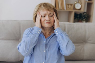 Evdeki kanepede oturan yaşlı bir kadının portresi. Baş ağrısı, ağrı ve kendini kötü hissetme ifadesi.