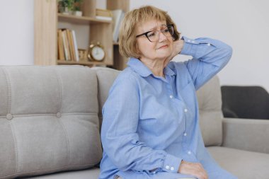 Yaşlı hasta yaşlı bir kadın kanepeye oturup boynuna dokunuyor. Omzu ve boynu ağrıyor. Sağlık ve Tıp Konsepti