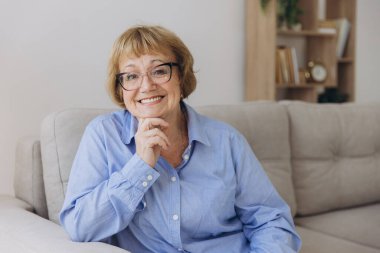 Gülümseyen, son sınıf sarışın, kameraya bakan mutlu, gözlüklü, evde poz veren yaşlı bir kadın oturma odasındaki portrede oturan, pozitif, bekar, emekli bir kadın.