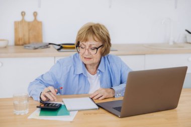 Hesap makinesini kullanarak kağıt fatura tutan kıdemli iş kadını, banka hesaplarını yöneten yaşlı kadın, bütçe vergisini hesaplayan, banka borcu emekliliği ödemesini planlayan ev mutfak masasında oturan yaşlı kadın..