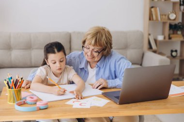 Mutlu büyükanne, kreş çocuğuna ev ödevi için yardım ediyor. Pozitif öğrenci kız ve büyükanne laptopta öğrenme kitabı okuyorlar, gülümsüyorlar, evde eğitimin tadını çıkarıyorlar.