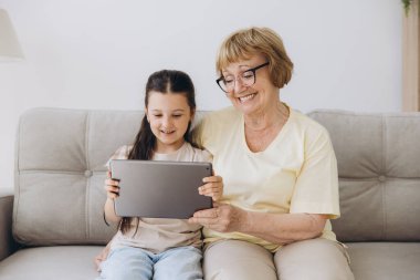 Eğleniyorum. Gülümseyen emekli kadın dadı rahat kanepeye oturup küçük kızı kucaklıyor internette tablet kullanarak komik çizgi filmler seyrediyor. Büyük anne ve küçük torun arasındaki bağ dijital bloknotta video klibinin keyfini çıkarın