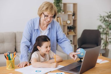 Mutlu büyükanne, kreş çocuğuna ev ödevi için yardım ediyor. Pozitif öğrenci kız ve büyükanne laptopta öğrenme kitabı okuyorlar, gülümsüyorlar, evde eğitimin tadını çıkarıyorlar.