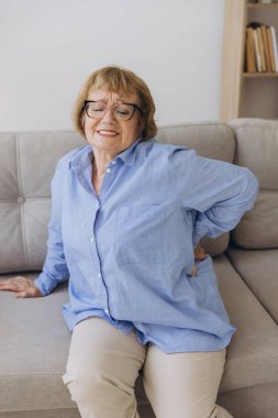 Mutsuz yaşlı bir kadın sırtına dokunuyor, acı çekiyor, oturma odasında kanepede oturuyor, sağlıksız orta yaşlı bir kadın sırt ağrısı çekiyor, omurga ağrısı çekiyor.