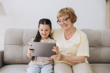 Eğleniyorum. Gülümseyen emekli kadın dadı rahat kanepeye oturup küçük kızı kucaklıyor internette tablet kullanarak komik çizgi filmler seyrediyor. Büyük anne ve küçük torun arasındaki bağ dijital bloknotta video klibinin keyfini çıkarın
