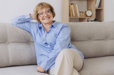 Gülümseyen, son sınıf sarışın, kameraya bakan mutlu, gözlüklü, evde poz veren yaşlı bir kadın oturma odasındaki portrede oturan, pozitif, bekar, emekli bir kadın.