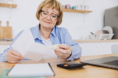 Hesap makinesini kullanarak kağıt fatura tutan kıdemli iş kadını, banka hesaplarını yöneten yaşlı kadın, bütçe vergisini hesaplayan, banka borcu emekliliği ödemesini planlayan ev mutfak masasında oturan yaşlı kadın..