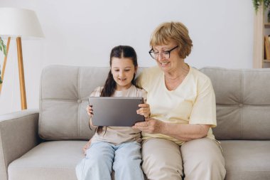 Eğleniyorum. Gülümseyen emekli kadın dadı rahat kanepeye oturup küçük kızı kucaklıyor internette tablet kullanarak komik çizgi filmler seyrediyor. Büyük anne ve küçük torun arasındaki bağ dijital bloknotta video klibinin keyfini çıkarın