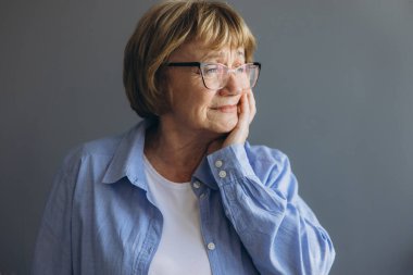 Yaşlı beyaz bir kadın evde oturuyor, yanağına dokunuyor ve acı içinde kıvranıyor. Şiddetli diş ağrısı ya da çene rahatsızlığı var gibi görünüyor..