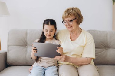 Eğleniyorum. Gülümseyen emekli kadın dadı rahat kanepeye oturup küçük kızı kucaklıyor internette tablet kullanarak komik çizgi filmler seyrediyor. Büyük anne ve küçük torun arasındaki bağ dijital bloknotta video klibinin keyfini çıkarın