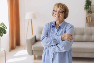 Gülümseyen gri saçlı kadın kameraya bakarak kollarını kavuşturdu. Evde poz veren mutlu yaşlı bayan. Oturma odasındaki portrede yaşlı, pozitif, emekli bir kadın.