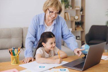Mutlu büyükanne, kreş çocuğuna ev ödevi için yardım ediyor. Pozitif öğrenci kız ve büyükanne laptopta öğrenme kitabı okuyorlar, gülümsüyorlar, evde eğitimin tadını çıkarıyorlar.