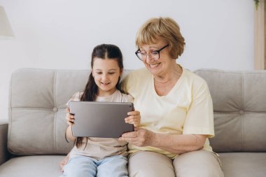 Eğleniyorum. Gülümseyen emekli kadın dadı rahat kanepeye oturup küçük kızı kucaklıyor internette tablet kullanarak komik çizgi filmler seyrediyor. Büyük anne ve küçük torun arasındaki bağ dijital bloknotta video klibinin keyfini çıkarın