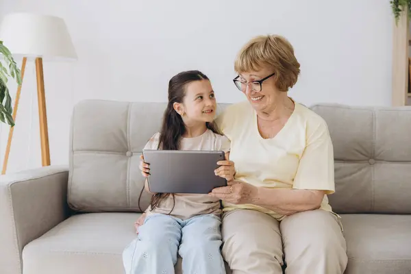 Stock image Having fun. Smiling retired woman nanny sit on cozy couch embrace little girl watch funny cartoon at web online using tablet. Bonding senior granny and small grandchild enjoy video clip on digital pad