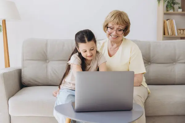 stock image Multi generational family, grandma and granddaughter sit on couch with wireless computer, look at each other with tenderness and love, enjoy leisure on internet and time together, watch on-line movies