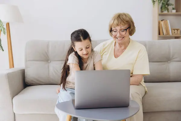 stock image Multi generational family, grandma and granddaughter sit on couch with wireless computer, look at each other with tenderness and love, enjoy leisure on internet and time together, watch on-line movies