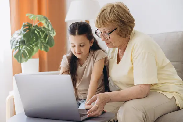 stock image Multi generational family, grandma and granddaughter sit on couch with wireless computer, look at each other with tenderness and love, enjoy leisure on internet and time together, watch on-line movies