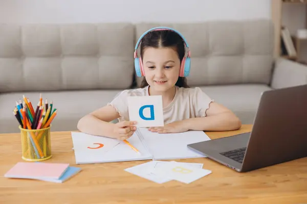 stock image Smiling small girl in headphones watch video lesson on computer with alphabet, happy little child in earphones have online web class using laptop at home, homeschooling concept