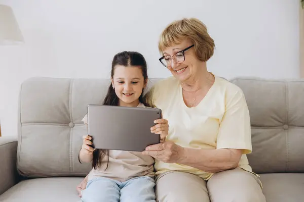 stock image Having fun. Smiling retired woman nanny sit on cozy couch embrace little girl watch funny cartoon at web online using tablet. Bonding senior granny and small grandchild enjoy video clip on digital pad