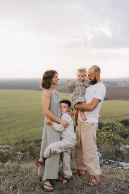 Dört kişilik neşeli bir aile açık havada manzaranın tadını çıkarıyor. Ebeveynler, çocuklarıyla birlikte, doğal bir ortamda sevgi ve mutluluk dolu bir kucaklaşmayı paylaşıyorlar..