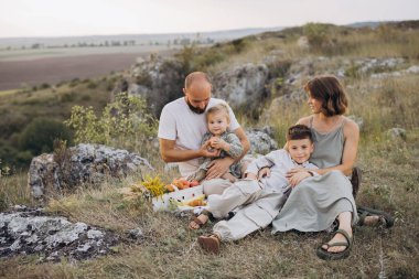 Resimli bir kırsal alanda piknik yapan neşeli bir aile. Ebeveynler ve çocuklar sıcak bir yaz gününde doğada birlikte iyi vakit geçiriyorlar..