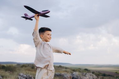 Neşeli genç bir çocuk, doğayla çevrili açık bir alanda dururken, özgürlüğü ve hayal gücünü sembolize eden oyuncak bir uçak tutuyor..