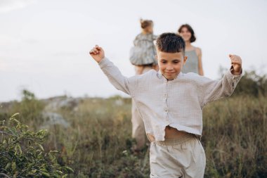 Mutlu bir çocuk çimenli bir alanda özgürce koşar, aile üyeleri arka planda, güneşli bir günün tadını çıkarırlar..
