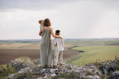 İki çocuğu olan bir anne kayalık bir tepede duruyor, geniş bir kırsal alana bakıyor. Sahne sükuneti, aile bağını ve doğanın güzelliğini yakalar..