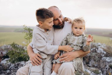 Bir baba, açık havada oğluyla ve kızıyla samimi bir anı paylaşıyor. Aile bağını ve sevgiyi simgeliyor..