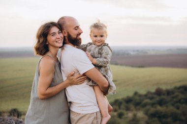 Üç kişilik gülümseyen aile, resimli manzaraya sevinçle sarılıyor. Ebeveynler ve çocuklar kırsalda güneşli bir günün tadını çıkarıyorlar, aşkı ve birlikteliği kutluyorlar..