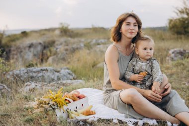 Bir anne ve bebek kırsal kesimde bir battaniyede birlikte oturuyorlar, bir piknik sepetiyle doğayla çevrili, sükunet ve bağlanma anının tadını çıkarıyorlar..