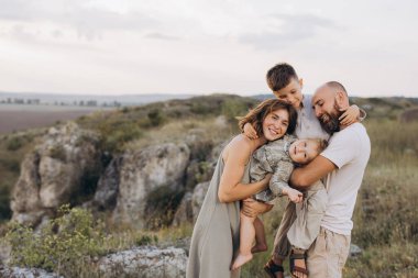 Dört kişilik neşeli bir aile, doğal bir ortamda kucaklaşıyorlar, mutluluk ve sevgi saçıyorlar. Ebeveynler ve çocuklar birlikte güzel bir günün tadını çıkarıyorlar..