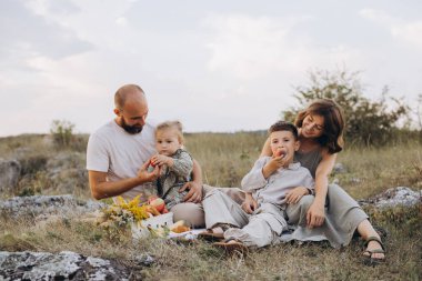 Neşeli bir aile piknikte, doğa tarafından çevrelenmiş, güzel bir tarlada taze meyve ve kahkahaların tadını çıkararak birlikte vakit geçiriyorlar..