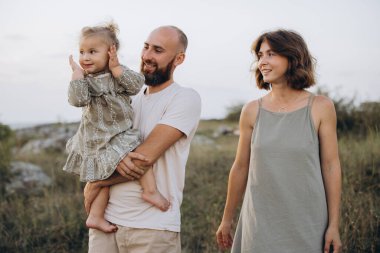 Üç kişilik neşeli bir aile, doğa manzaralı bir yerde neşeli bir anı paylaşıyor. Sevgi ve birlikteliği vurguluyor..