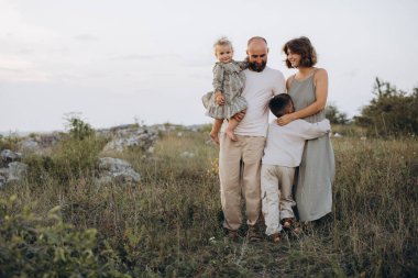 Açık havada dört kişilik bir aile, sakin bir manzarada sevgi ve beraberlik sergiliyor. İki çocuklu ebeveynler, doğanın güzelliğinin ortasında, yumuşak bir akşam gökyüzünün altında huzurlu anların tadını çıkarıyorlar..