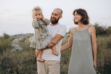 Dışarıda huzurlu bir günün tadını çıkaran, babası, annesi ve çocuğu olan neşeli bir aile. Ebeveynler genç kızlarına sevgiyle sarılırken doğal bir ortamda gülümsüyorlar..