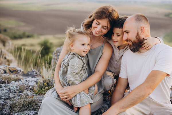 Радостная семья из четырех человек обнимает друг друга, сидя на скалах в естественной обстановке, иллюстрируя любовь, счастье и единение..