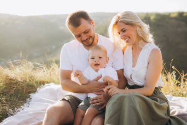 Üç kişilik neşeli bir aile birlikte dışarıda vakit geçiriyorlar. Gülümseyen ebeveynler ve bebekleri piknikte güneşli güzel bir günün tadını çıkarıyorlar..