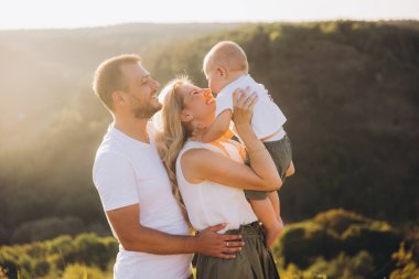 Güneşli bir açık havada kucaklaşan ve gülümseyen üç kişilik mutlu bir aile. Ebeveynler ve çocuklar birlikte kaliteli zaman geçiriyorlar, doğada bir neşe ve beraberlik anı yakalıyorlar..