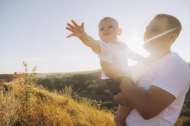 Bir baba bebeğini neşe içinde güneşli bir tepede tutar, sıcak ve sevgi dolu bir anı yakalar..