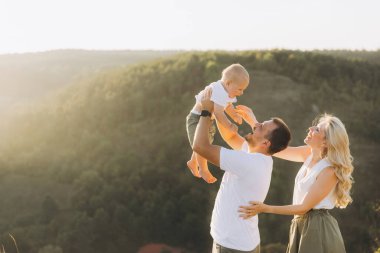 Neşeli bir aile açık havada güneşli bir günün keyfini çıkarırken ebeveynler neşeli bebeklerini güzel doğal bir zemine doğru kaldırıyorlar..